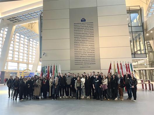 Die Exkursionsgruppe lässt sich vor einer riesigen Tafel im Inneren der EZB ablichten. Auf dieser Tafel ist der Name der Europäischen Zentralbank in allen Sprachen der EU zu lesen.